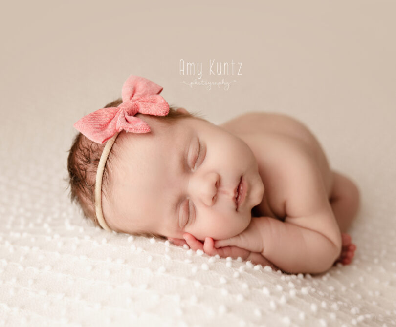 newborn baby sleeping peacfully during her newborn photoshoot. She is wearing a pink bow and you can see her fingers and toes.