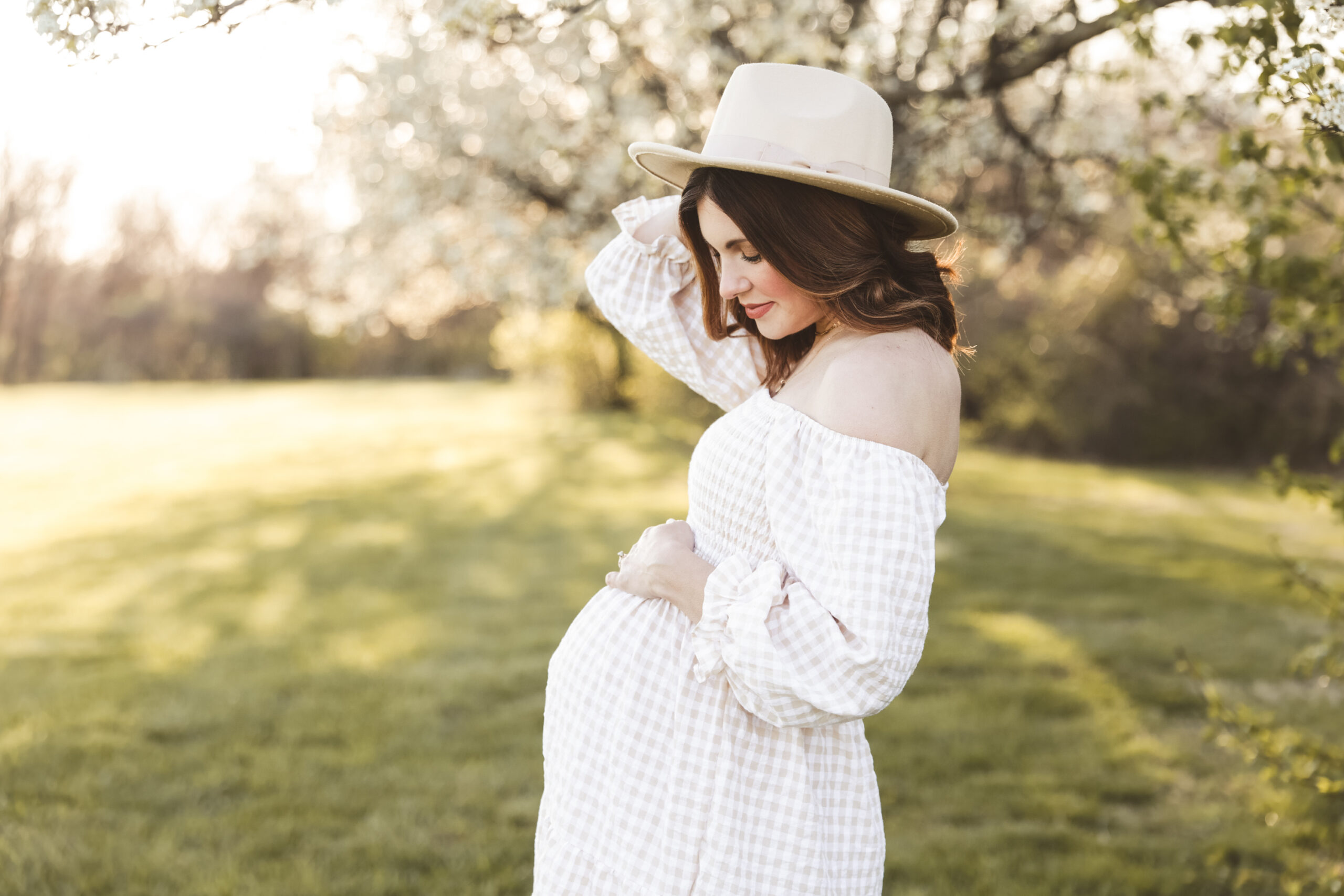 a pregnant mother standing infront of white flowering trees, holding her hat and looking lovingly at her baby bump